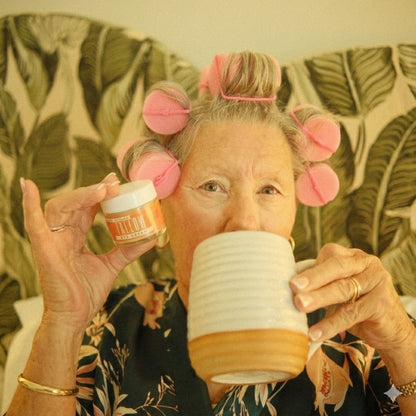 Person with pink hair rollers holding a jar and a mug in front of a patterned background